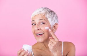 smiling beautiful mature woman holding jar of moisturizer and looking at camera over pink background