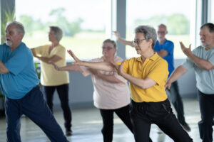 A small group of seniors are seen practicing Tai Chi in an indoor studio. They are each dressed comfortably and are focused on their movements and proper form as they practice the Martial Art.
