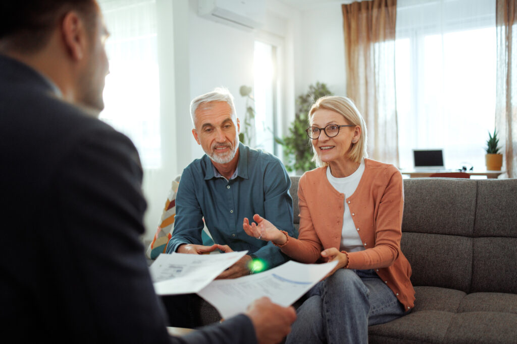 Mature couple sitting in their living room, engaged in a friendly conversation with a professional insurance agent. They are discussing documents, showing mutual interest and understanding in a relaxed, homey atmosphere.